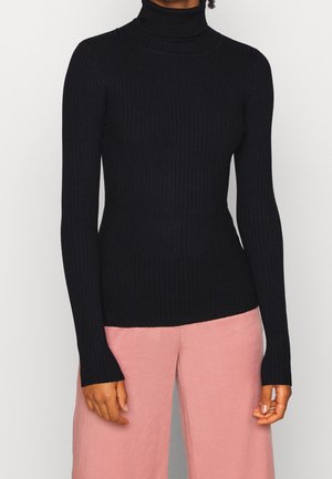 Kvinde iført en tætsiddende sort ribstrikket rullekravetrøje med lange ærmer og løse lyserøde bukser, stående mod en lys baggrund.