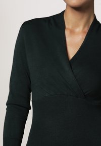 Sort, langærmet bluse med V-hals design. Har en tætsiddende krop og ribbet tekstur omkring taljen. Blødt stof, minimal detaljering.