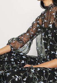 Sort gennemsigtig kjole med broderede blå blomsterpatroner, høj hals og rynkede ærmer. Har en flydende silhuet og tekstureret stof.