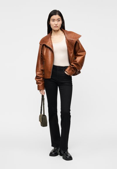 Brun läderjacka med bred krage, kombinerad med en beige topp, svarta jeans och klumpiga svarta skor. Modellen håller en grön väska.