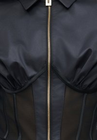 Gros plan sur un corset noir avec des bonnets structurés, des panneaux en maille et une fermeture éclair centrale dorée sous un col.