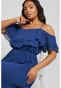 Ljusblå klänning med avskurna axlar i lätt tyg, som har lager av volanger, elastisk midja och en mjuk, flödande silhuett.