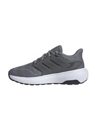 Zapatilla deportiva gris con parte superior de malla, tres franjas prominentes, suela blanca acolchada y suela negra. Presenta un sistema de cordones y un collar acolchado.