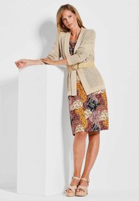 Beiger Netz-Cardigan über einem multicolor gestreiften Kleid in verschiedenen Orangetönen, Burgunderrot und Schwarz. Frau trägt beiger Plateausandaletten.