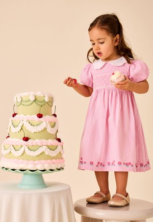 Noor tüdruk roosas kleidis seisab pingi peal, hoiab käes muffin’t ja kirssi ning vaatab suurt pastelse rohe-roosa korruselist tordi.