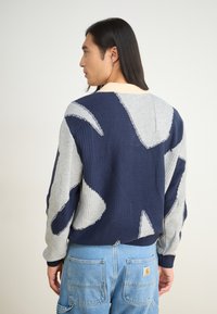 Πλεκόμενη μπλούζα με ναυτικό και γκρι γεωμετρικό σχέδιο, ριμπ μανίκια και στρογγυλή λαιμόκοψη, φορεμένη πάνω από ανοιχτό μπλε cargo παντελόνι.