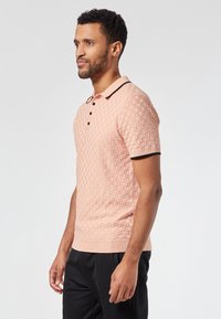 Lyhythihainen polo-paita, jossa on teksturoitu vaaleanpunainen neulos, mustalla viimeistellyt kaulus, kolme nappia ja ribbaus helma.