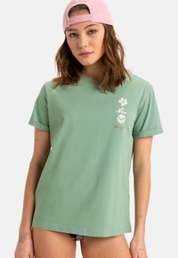 Grøn bomulds T-shirt med optrækning af ærmerne, prydet med blomster grafik og teksten "Roxy" i en kontrastfarve på forsiden.