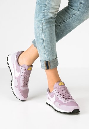 Baskets Nike violettes avec le logo blanc, tissu texturé, bout arrondi et semelle coussinée ; portées avec un jean bleu clair retroussé.
