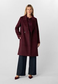 Cappotto in lana borgogna con design doppiopetto, ampi risvolti, tasche laterali, abbinato a pantaloni in denim scuri a gamba larga e scarpe coordinate.