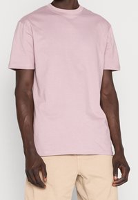 T-shirt en coton rose clair à manches courtes et col rond, avec une texture lisse et une coupe décontractée, associé à un pantalon beige.