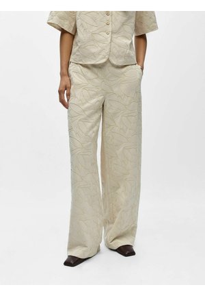 Krémové nohavice s širokými nohavicami a textúrovaným listovým vzorom, nosené s rovnako farebnou bundou s polovičnými rukávmi a gombíkmi, doplnené tmavými topánkami so štvorcovou špičkou.