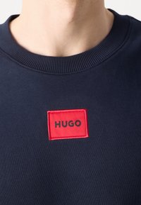Sweat-shirt bleu marine à col rond avec une écusson rectangulaire rouge affichant le nom de la marque "HUGO" en lettres noires sur la poitrine.