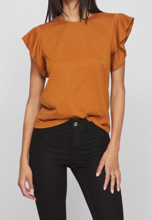 Vrouw draagt een roestoranje top met ruches aan de mouwen, lichtjes ingestopt in een zwarte, hoog uitgesneden jeans, staand met één hand rustend nabij haar dij.