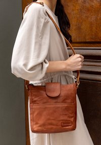 Brun läder crossbody-väska med flap stängning, med en framficka och justerbart axelband. Mjuk textur och rektangulär form.