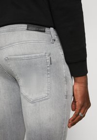 Jeans de mezclilla gris con una textura suave, que presentan un diseño de bolsillo trasero y una etiqueta de cuero negra cerca de la cinturilla. El conjunto incluye una blusa negra.