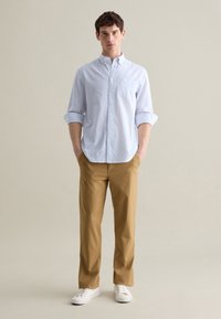 Camisa de botones a rayas azul claro con mangas enrolladas, combinada con chinos beiges y zapatillas blancas, de pie contra un fondo neutro.
