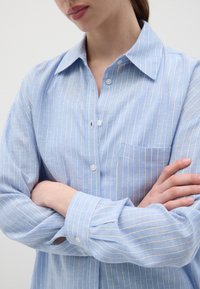 Camicia azzurra chiaro con sottili righe verticali e una tasca sul petto. Presenta un colletto classico e maniche lunghe e rilassate.