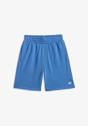 Garçon portant un t-shirt vert « Fair Play » et un short bleu, debout avec un pied sur un ballon de football blanc sur fond gris uni.