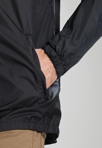 Czarna, lekka kurtka z elastycznymi mankietami, bocznymi kieszeniami i gładką, lekko błyszczącą fakturą. Dół jest prosty, z minimalnymi detalami projektowymi.