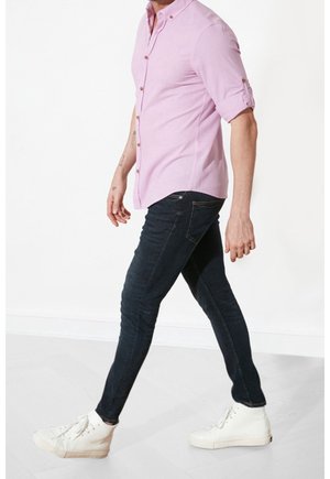 Uomo che cammina indossando una camicia rosa chiaro con bottoni e maniche arrotolate, jeans skinny scuri e sneakers alte bianche.