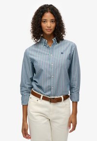 Koszula z button-up w niebiesko-zielone paski, wykonana z bawełny, z długimi rękawami i kołnierzykiem, z małym granatowym logo na piersi. Połączona z jasnymi spodniami.