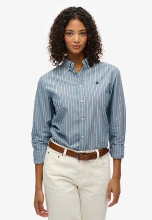 Camisa abotonada a rayas azul y verde, de material de algodón, de manga larga, con cuello, con un pequeño logo azul marino en el pecho. Combinada con pantalones claros.