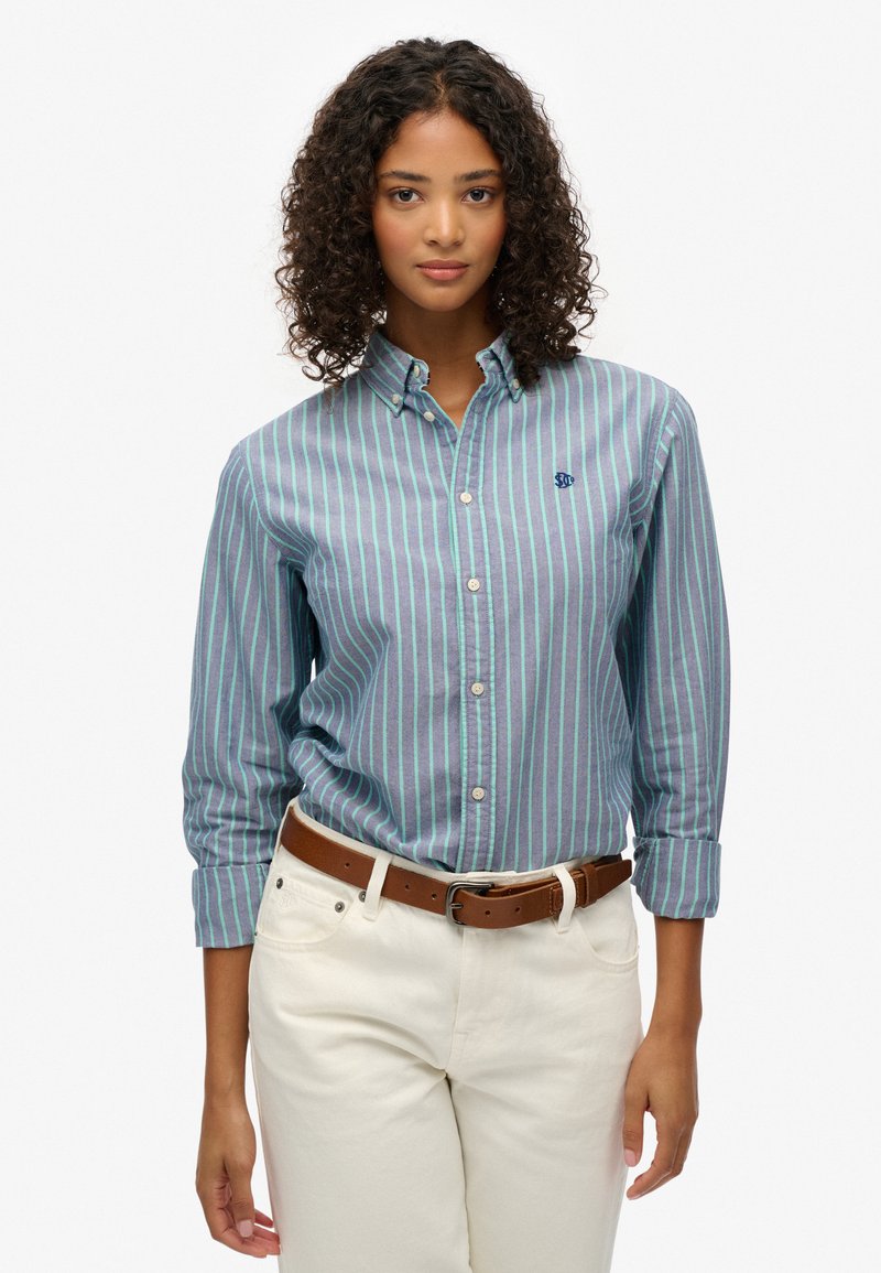 Koszula z button-up w niebiesko-zielone paski, wykonana z bawełny, z długimi rękawami i kołnierzykiem, z małym granatowym logo na piersi. Połączona z jasnymi spodniami.