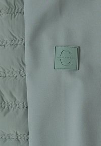 Lähikuva tikatuista ja sileistä harmaista kangasosista, jossa on tikattu neliönmuotoinen merkki, jossa lukee "C OUTERWEAR" sileässä kankaassa.