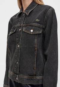 Femme portant une veste en jean noire avec des poches poitrine boutonnées et un petit logo crocodile vert sur le côté gauche.