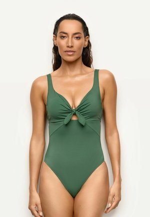 Mujer con el pelo mojado, con un traje de baño verde de una pieza con detalle de atado en la parte delantera, de pie frente a un fondo claro y liso.