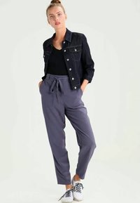 Moteris stovi su rankomis kišenėse, vilkinti tamsiai džinsinę striukę, juodą marškinėlį, pilkas kelnes su juostele ir baltus sportbačius ant vientiso fono.