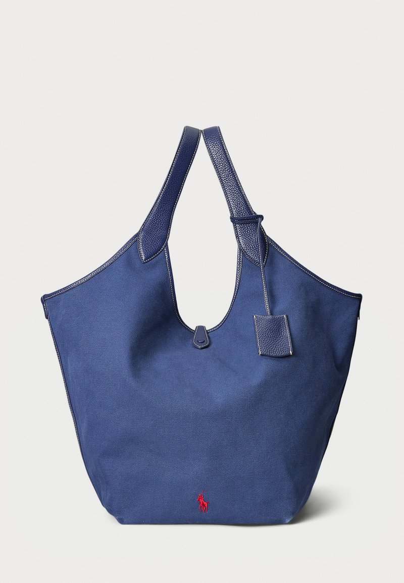Mörkblå canvas toteväska med en strukturerad form, utrustad med läderdetaljer, dubbla handtag och en liten fäst tag. Röd logobeskrivning.
