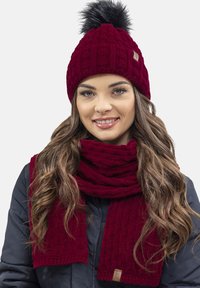 Berretto lavorato a maglia bordeaux con pompon nero e sciarpa coordinata, caratterizzato da una texture a coste e un piccolo etichetta con logo.
