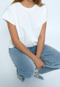 Bílé tričko s krátkým rukávem a volným střihem, kombinované se světle modrými džínami a metalickými balerínami se špičatou špičkou a cvočky.