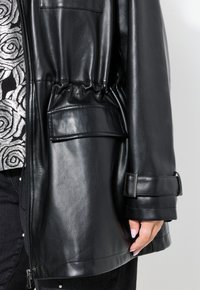 Giubbotto in ecopelle nera con vita elasticizzata, tasca a toppa frontale e dettagli con cinturini sui polsini. Texture liscia e design elegante.