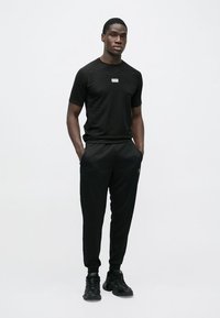 Melns sporta tērps ar īsām piedurknēm un sašaurinātām biksēm. Krekla priekšā redzams logotips, gluda tekstūra un pieguļošs dizains.