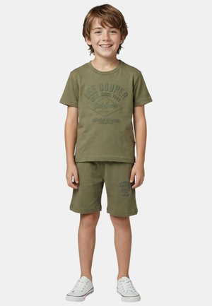 Garçon souriant portant un t-shirt Lee Cooper vert olive et un short assorti, avec des baskets blanches, debout devant un fond uni.