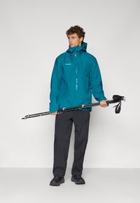 Turquoise waterproof jacket with a hood, zip closure, and logo; paired with dark pants and holding trekking poles.