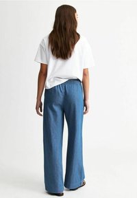 Bílý tričko s krátkým rukávem v kombinaci s džínovými kalhotami širokého střihu, které mají elastický pas a světlou modrou barvu. Hladká textura, minimalistický design.