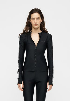 Žena má na sebe priliehavú čiernu bundu na zips a zodpovedajúce legíny s logom „Moschino“ na rukávoch na jednoduchom svetlom pozadí.