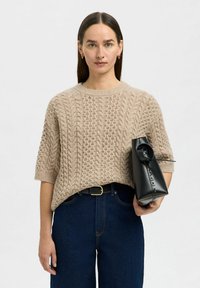Suéter de punto beige con un patrón texturizado, mangas cortas, usado sobre unos vaqueros de mezclilla azul oscuro. Sosteniendo un bolso estructurado negro.