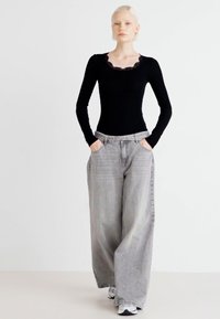 Top preto de manga longa com decote bordeado de renda; jeans de perna larga cinza claro; modelo de pé com as mãos nos bolsos; tênis.