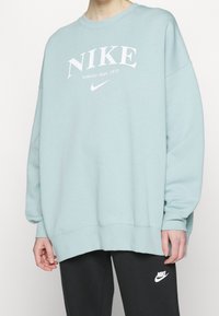 Světlomodrá oversized mikina Nike s bílým tištěným logem a textem. Má žebrované manžety a lem, měkkou texturu a uvolněný střih.