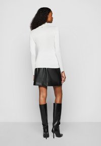 Weißes Langarmshirt mit leichtem Glanz, kombiniert mit einem schwarzen Leder-Minirock und kniehohen schwarzen Stiefeln mit Absatz.