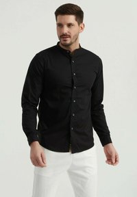 Chemise noire à manches longues avec col montant, au toucher lisse; dotée de boutons bleus et d'une touche de tissu coloré à l'ourlet.