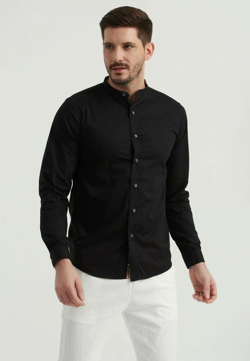 Chemise noire à manches longues avec col montant, au toucher lisse; dotée de boutons bleus et d'une touche de tissu coloré à l'ourlet.