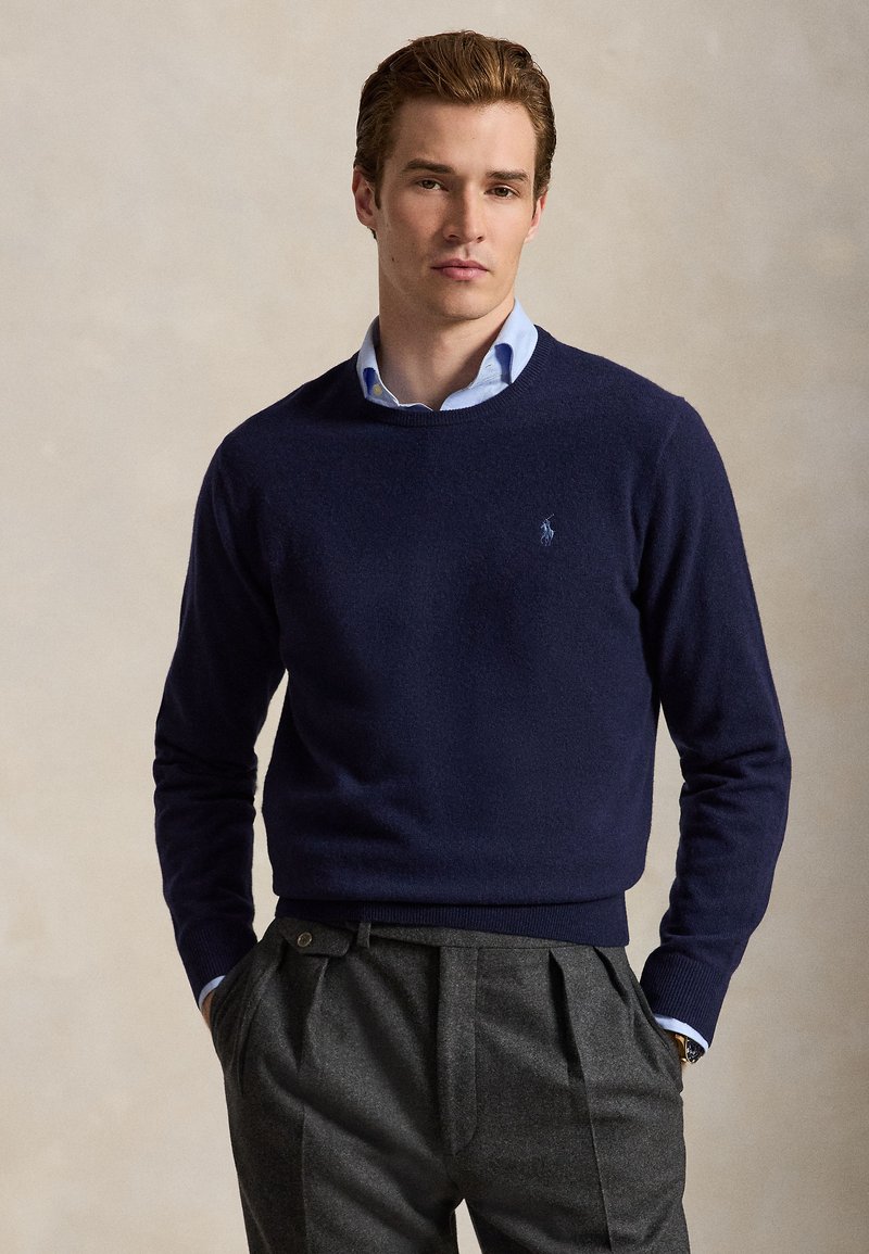 Ung mand iført en mørkeblå sweater over en lyseblå skjorte med krave og grå plisserede bukser, stående med hænderne i lommen.
