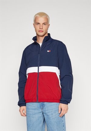 Chaqueta cortavientos azul marino y roja con una franja horizontal blanca. Presenta un cuello alto y puños elásticos, fabricada con un tejido ligero.