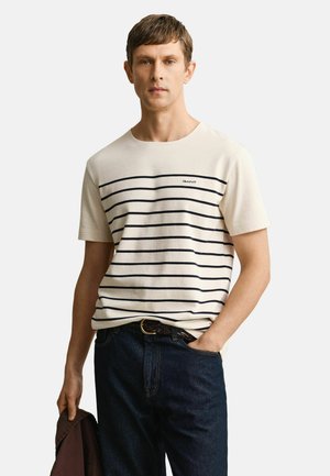 Mann trägt weißes Kurzarmhemd mit marineblauen horizontalen Streifen und dunkle Jeans, hält eine gefaltete braune Jacke in der linken Hand.